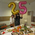 Dwie kobiety widoczne na zdjęciu. Jedna trzyma w ręku balony w kształcie cyfry 2 i 5. Na stole widoczne jedzenie