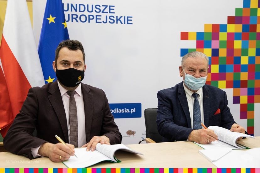 Dwóch mężczyzn w garniturach siedzi za biurkiem i podpisuje dokumenty na tle flagi UE
