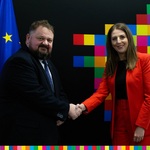 Mężczyzna ściska dłoń kobiecie na tle czarne ściany z kolorowymi kwadratami oraz flagą UE w tle