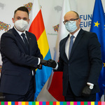 Dwóch mężczyzn w garniturach ściska sobie na wzajem dłoń. W tle flaga województwa podlaskiego, Polski oraz UE