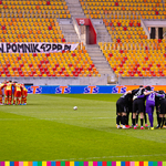 Piłkarze dwóch drużyn nachyleni nad sobą. Po prawej w czarnych strojach, po lewej w żółto-czerwonych.