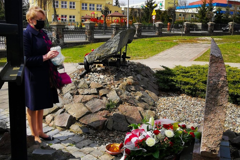 Kobieta trzyma znicz stojąc przed tablicą pamiątkową. Przed nią leżą kwiaty. W tle miejskie zabudowania.