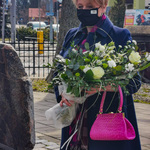 Wiesława Burnos z kwiatami w masce na twarzy stojąca przed tablicą pamiątkową w Sokółce 
