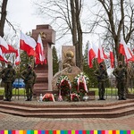Widoczny pomnik. Wokół niego powiewające biało-czerwone flagi oraz żołnierze trzymający wartę honorową