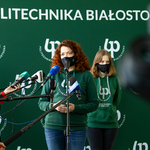 Trzy kobiety stoją przed mikrofonami. W tle ścianka z napisem 