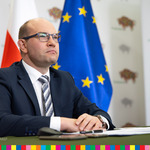 Marszałek Artur Kosicki siedzi za stołem w tle flagi województwa podlaskiego, Polski oraz Unii Europejskiej