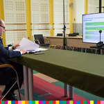 Marszałek siedzi przy stole nakrytym zielonym obrusem i przegląda dokumenty. W tle widoczny monitor z wyświetlonym slajdem oraz kamera