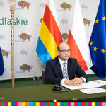 Marszałek Artur Kosicki siedzi za stołem z dokumentami. W tle stoi flaga województwa podlaskiego, Polski i UE oraz laska marszałkowska z orłem