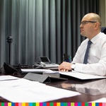 Mężczyzna w białej koszuli pod krawatem. Na biurku widoczne dokumenty. Z lewej storny widoczny monitor oraz stojąca kamera internetowa