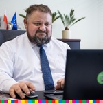 Mężczyzna w koszuli pod krawatem siedzący za komputerem. W tle widoczne trzy flagi Polski, UE, Województwa Podlaskiego oraz doniczka.