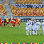 Piłkarze obu drużyn stoją na boisku w dwóch grupach. W tle widoczne trybuny