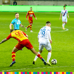 Piłkarz Jagielloni Białystok próbuje nogą zabrać piłkę przeciwnikowi. W tle widoczny sędzia i kilku piłkarzy