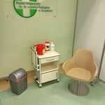 Fotel, półka z rzeczami oraz kosz na śmieci. Na ścianie wisi logotyp suwalskiego szpitala
