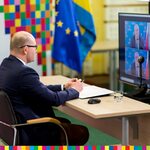 Marszałek Artur Kosicki siedzący za stołem.Przed marszałkiem znajduje się ekran z widoczna wideorozmową. W tle widoczne są flagi województwa, Unii Europejskiej oraz ścianka  z logotypami regionu.