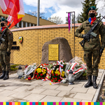 Kamień z tablicą: Koszary 10 Pułku Ułanów Litewskich. Pod nim leżą wiązanki z kwiatów. Obok kamienia z tablicą pamiątkowa stoi dwóch żołnierzy