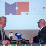 Dwóch mężczyzn jest odwróconych w stronę ekranu przedstawiającego logotyp Muzeum Podlaskiego