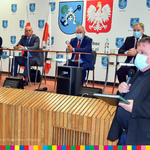 Mężczyzna stojący z mikrofonem w dłoni podczas XXVIII sesji Rady Miejskiej w Jedwabnem. Za nim przy stolikach siedzi czterech mężczyzn.