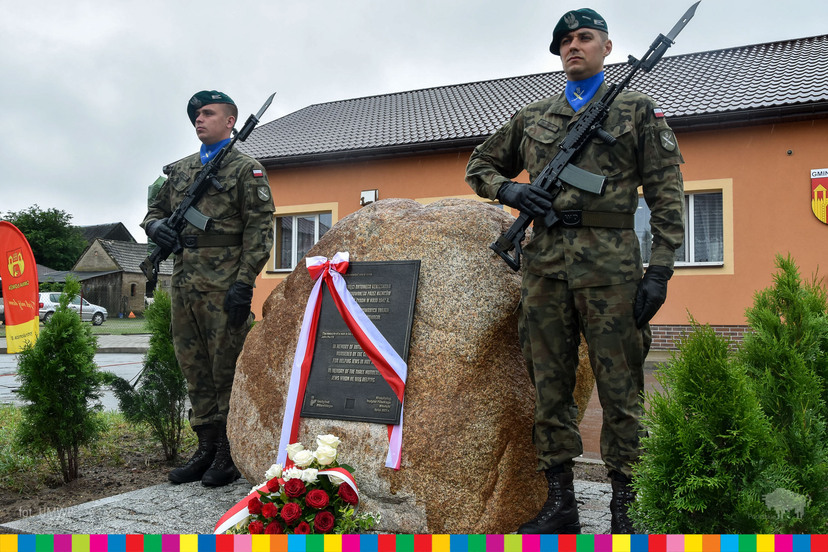 Dwóch żołnierzy stojących przy głazie z tablicą pamiątkową. Tablica przewiązana biało-czerwoną wstęgą