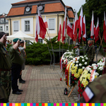 Umundurowani mężczyźni salutują. Widoczne kolorowe wieńce oraz flagi biało-czerwone
