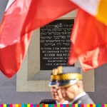 Tablica wisząca na murze. Widoczne powiewające flagi oraz głowy mundurowych w czapkach
