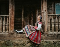 Dziewczyna ubrana w strój ludowy siedząca na ganku drewnianego domu