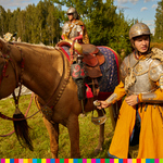 Mężczyzna ubrany w zbroję, hełm, sukno, skórę stoi obok konia i trzyma w ręku strzemię