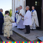 Przed wejściem do kościoła stoi trzech kapłanów i mężczyzna ubrany w białą komżę trzymający kropidło i naczynie z wodą święconą. Przed nimi stoi kilka osób na schodach kościoła