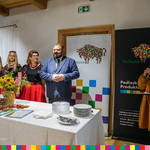 Mężczyzna stoi za stołem z talerzami i kwiatami obok czterech kobiet ubranych w stroje ludowe. Po prawej stoi kobieta z mikrofonem
