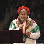 Starsza kobieta w wianku z kwiatów na głowie trzyma w ręku mikrofon