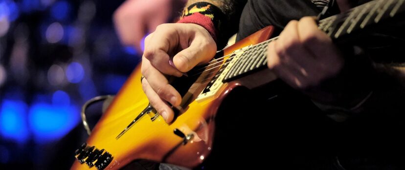 Zbliżenie na gitarę elektryczną, na której gra mężczyzna, dłonie z kostką nad strunami gitary