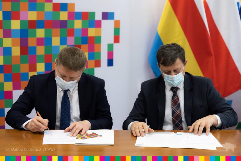 Dwóch mężczyzn podpisuje dokumenty. W tle widać flagę niebiesko-żółto-czerwono-białą