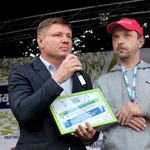 członek zarządu Marek Malinowski trzyma mikrofon, w drugiej podziękowania, obok niego stoi mężczyzna w czerwonej czapce z napisem Organizator