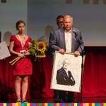 Aktor Jerzy Stuhr przy mikrofonie w rękach trzyma portret Andrzeja Wajdy. Za nim pomiędzy dwoma mężczyznami stoi kobieta w czerwonej sukience w rękach trzyma żółte słoneczniki