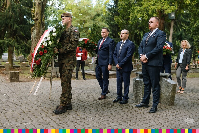 Żołnierz trzyma wieniec z kwiatów biało-czerwonych, za nim stoją Artur Kosicki, Sebastian Łukaszewicz, inni uczestnicy wydarzenia.