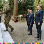 Żołnierz trzyma w dłoniach wieniec z kwiatami, za nim stoją marszałek Kosicki, wicemarszałek Łukaszewicz i radny Perkowski.