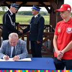 Mężczyzna podpisuje dokument przy stole, z anim mężczyźni w mundurach, po prawej stronie chłopak ubrany na czerwono 