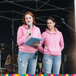 Dwie kobiety ubrane w różowe bluzy, jedna z nich trzyma przy ustach mikrofon.