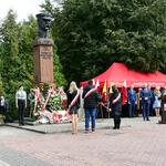 Przed pomnikiem stoi troje ludzi przewiązanymi wstęgami biało-czerwonymi, po prawej stronie uczestnicy wydarzenia.